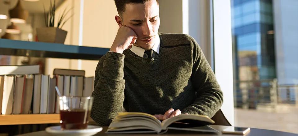 Ein nachdenklicher Mann in einem grünen Pullover sitzt an einem Tisch in einem modernen Café, liest ein Buch und stützt seinen Kopf auf eine Hand. Neben ihm steht ein Glas Tee auf einem Untersetzer, und im Hintergrund sind Bücherregale und Pflanzen zu sehen. Durch das Fenster fällt natürliches Licht, das die ruhige und konzentrierte Atmosphäre unterstreicht.