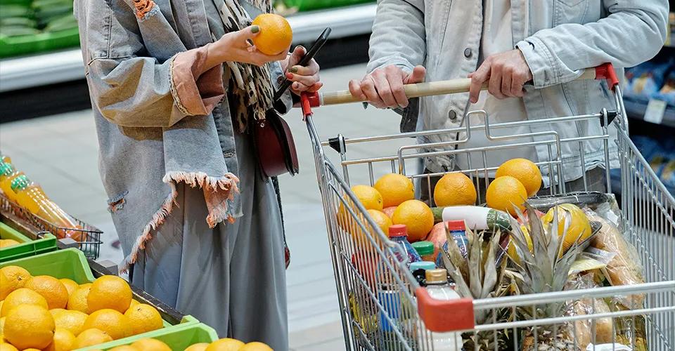 Zwei Personen stehen in einem Supermarkt, eine hält eine Orange in der Hand, während die andere einen Einkaufswagen schiebt, der mit Orangen, Ananas und verschiedenen Lebensmitteln gefüllt ist. Beide tragen lässige Kleidung, und im Hintergrund sind weitere Obstkisten und grüne Produkte zu sehen. Die Szene vermittelt den Eindruck eines normalen Einkaufsalltags.