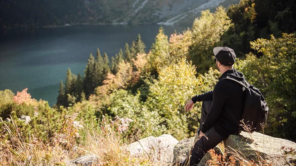 Ein Mann mit schwarzer Kappe und Rucksack sitzt auf einem Felsen und blickt auf einen See inmitten von Bergen und dichten Wäldern. Es ist Herbst, und die Bäume zeigen eine Mischung aus grünen, gelben und roten Blättern. Der Mann geniesst die Aussicht auf die ruhige, natürliche Landschaft unter klarem Himmel.