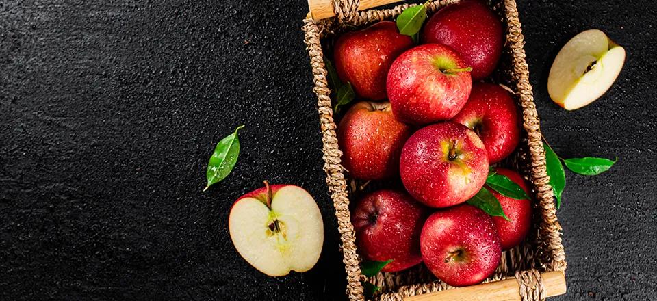 Ein Korb voller reifer, roter Äpfel, einige mit grünen Blättern, steht auf einem dunklen, strukturierten Untergrund. Neben dem Korb liegen zwei Apfelhälften und ein einzelnes grünes Blatt. Die Äpfel sind mit Wassertropfen bedeckt, was Frische symbolisiert.