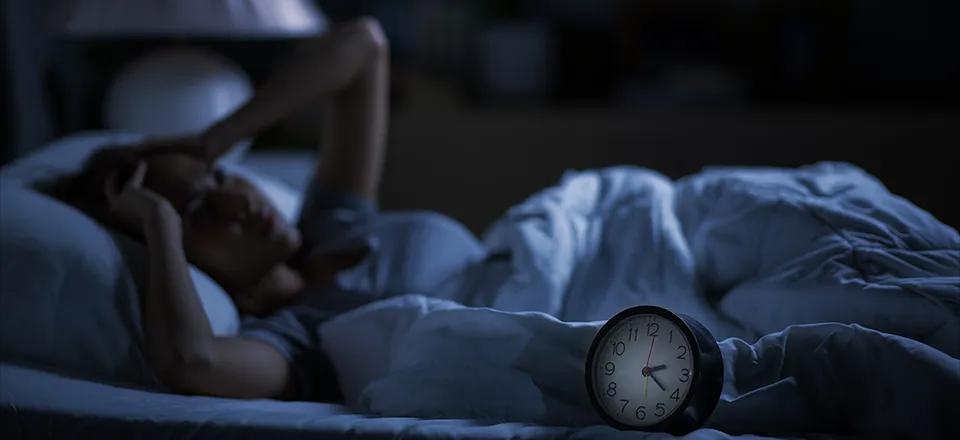 Eine Frau liegt nachts schlaflos im Bett, hält sich den Kopf und schaut nachdenklich. Eine Uhr auf dem Nachttisch zeigt 2:30 Uhr an, das Zimmer ist dunkel beleuchtet.